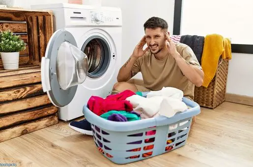 man putting dirty laundry into washing machine covering ears with fingers with annoyed expression for the noise of loud music